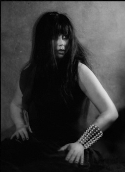 Long haired woman sitting with hands on her legs. She wears a wide studded bracelet on one forearm.